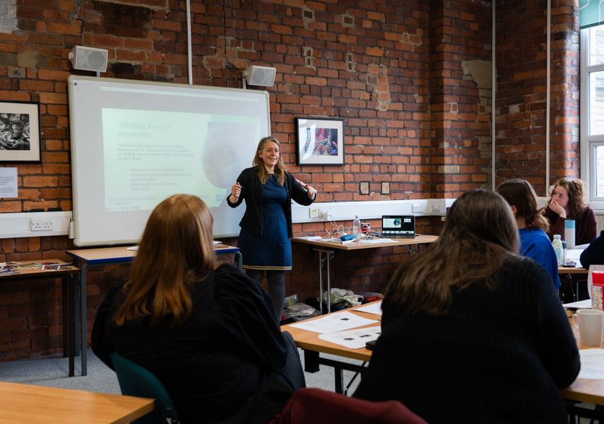 A woman stood in front of people in a brick walled room. She seems dynamic and trying to engage with everyone.