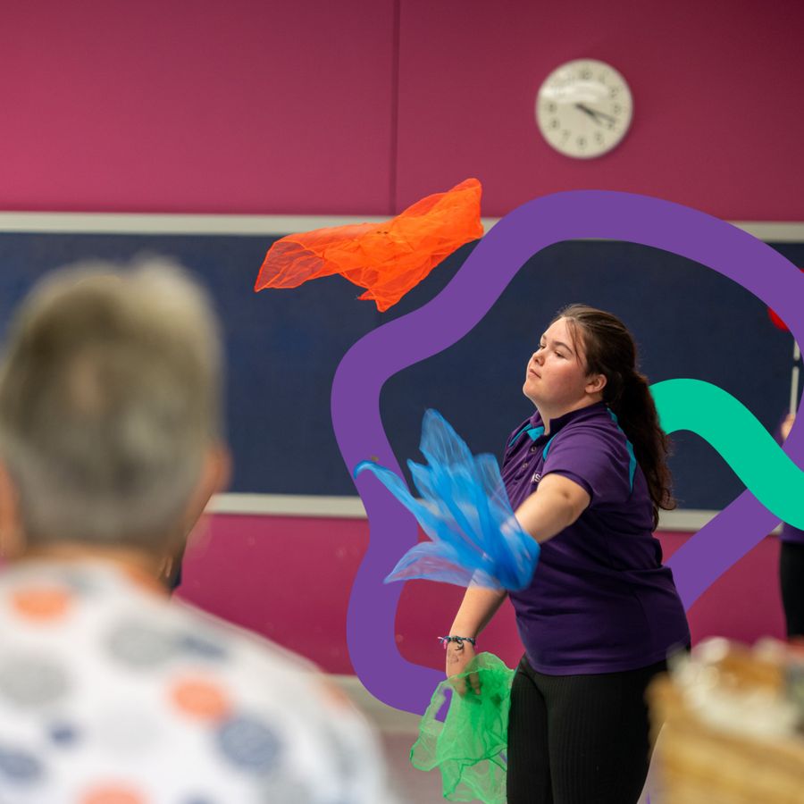 A young girl in a school is juggling colourful cloths. There are other children in the background.