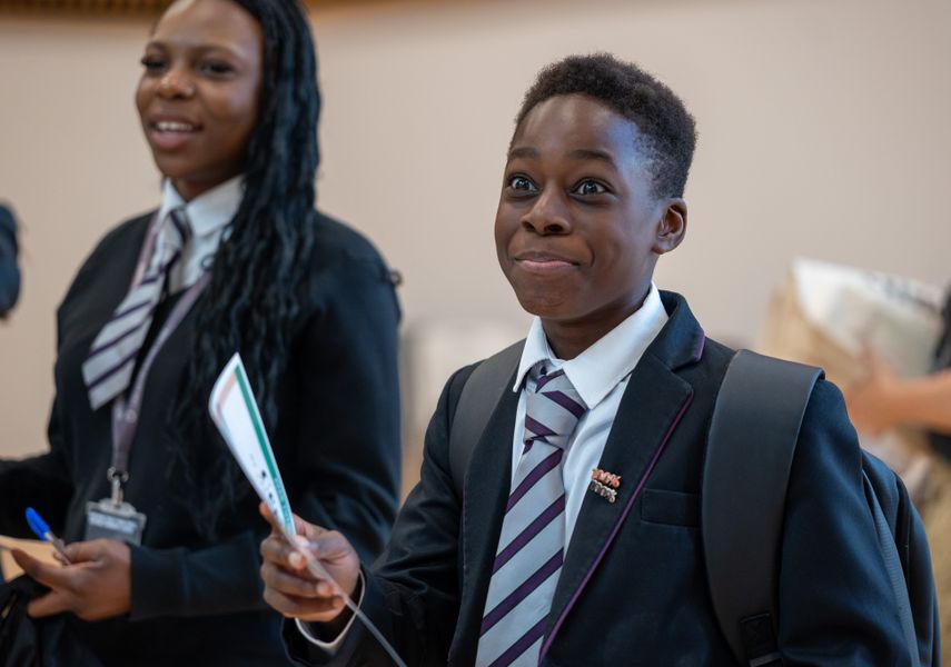 A young boy in a school uniform smiling and holding a piece of paper. A girl in the back is also smiling.