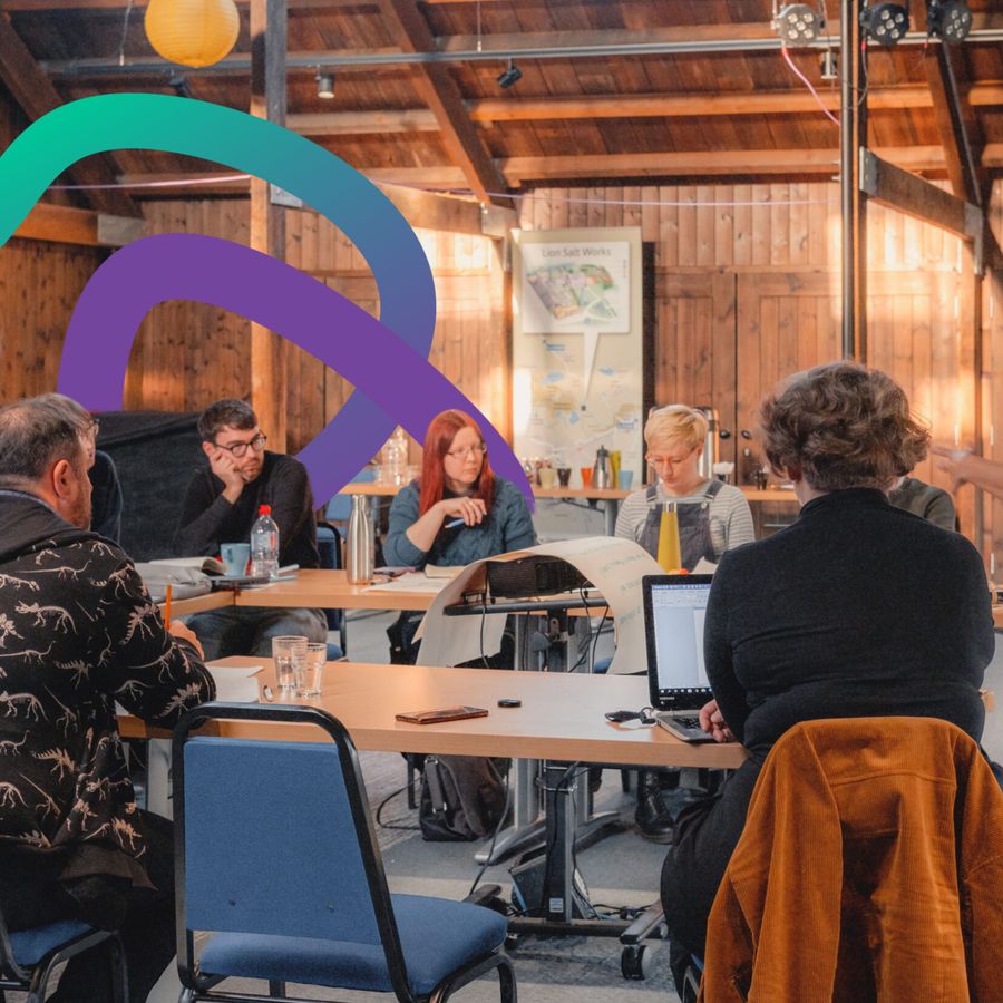 A group of cultural practitioners are attending a workshop. They are sat around a table and look deep in concentration. A workshop leader is stood to the right of the image, presenting to them.