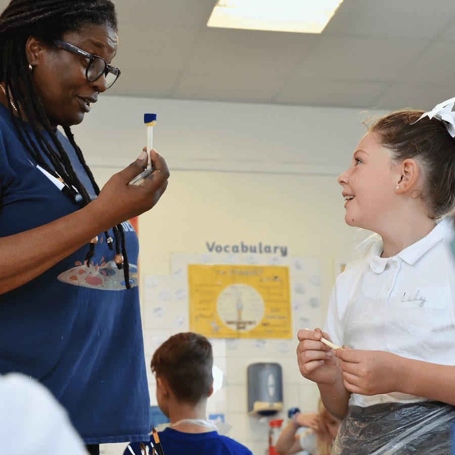 A pupil engages with a visiting teaching artist in a school setting. They are looking directly at each other. The artist is holding up a paint brush to show the pupil, who is grinning at her. 
