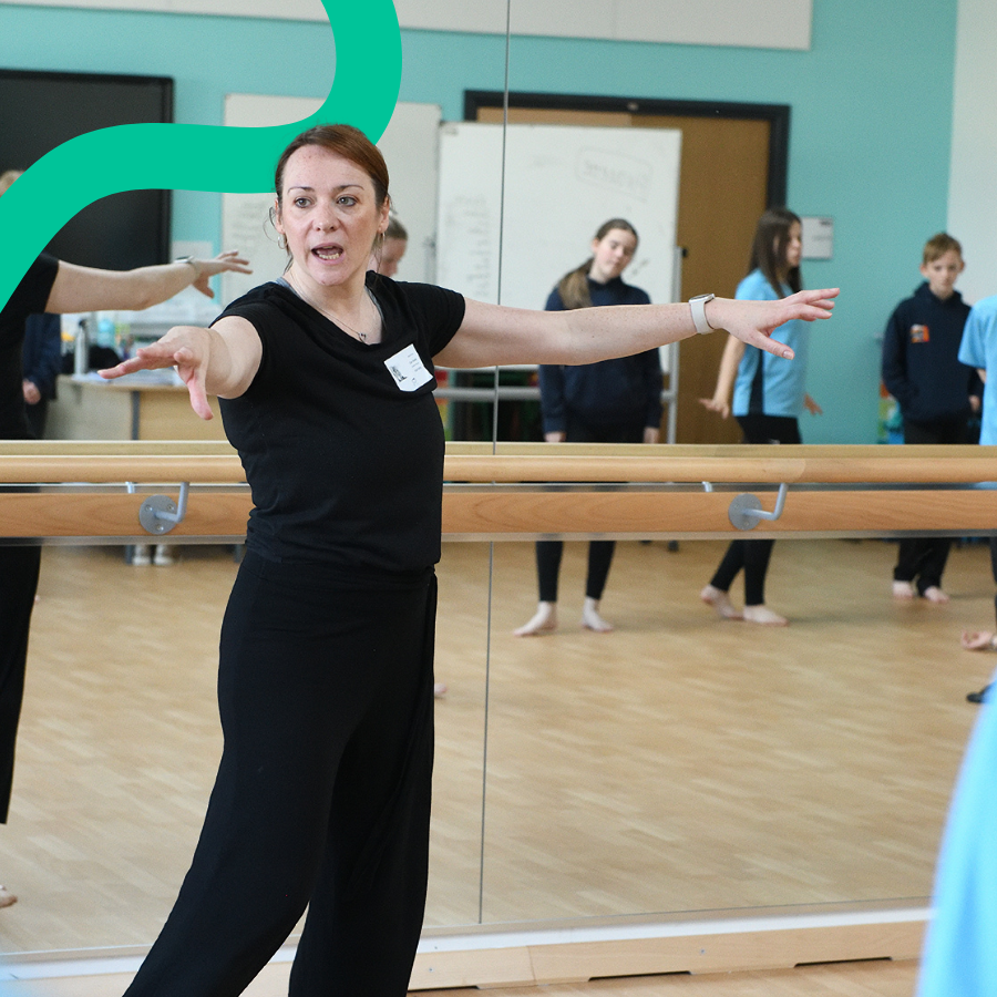 A drama teacher dressed in black is demonstrating a move to students, with her arms outstretched. She is stood in front of a mirror with a dance bar. The reflection of the students is in the mirror.