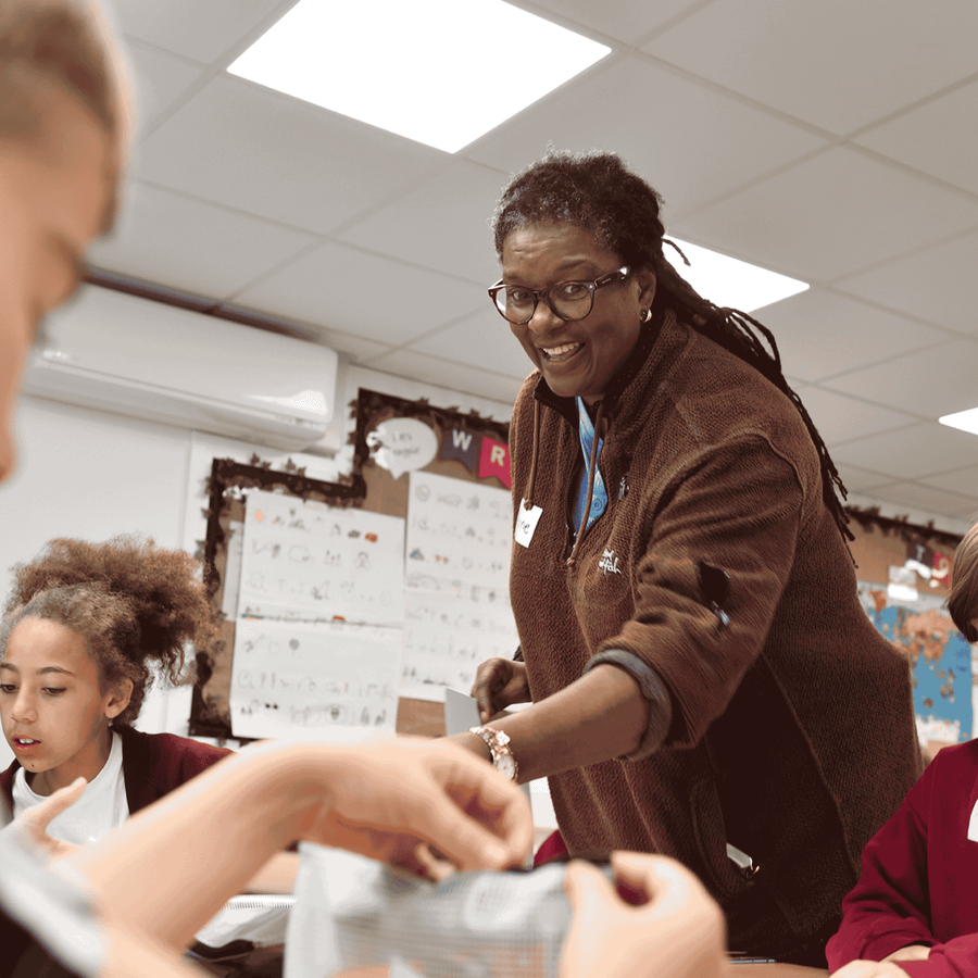 A teacher stood reaches down, with a smile on their face, to help a young student. 