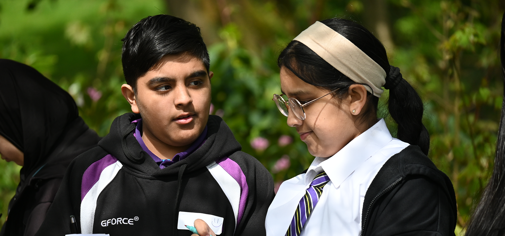 Two Falinge Park High Schools compare notes whilst working on a project. They are outside, surrounded by foliage. The sun is shining.